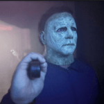 A person wearing a distressed, eerie mask holds a small object in a dimly lit room with a misty atmosphere Their expression is obscured by the mask, adding to the unsettling mood